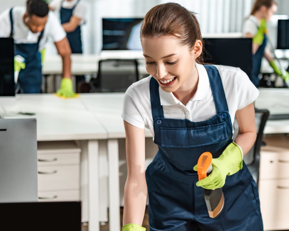 cheerful cleaner in overalls cleaning office desk with rag